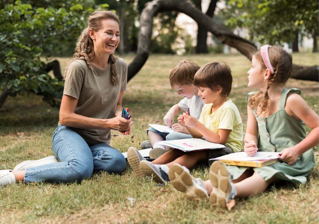 Woman sitting on grass with three children, all holding books and pencils Toptestbank